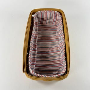 May include: A rectangular woven wood basket with a light tan finish and a removable fabric liner. The liner features a repeating pattern of thin vertical stripes in shades of red, blue, and white, covering the interior base and sides.