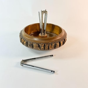 May include: A round wooden bowl with a natural bark edge and a central metal stand holding three silver-toned nutcrackers. An additional matching silver-toned nutcracker is positioned separately in front of the bowl on a flat surface.