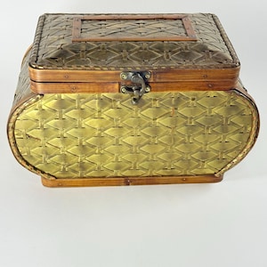 May include: A rectangular storage box with a woven metallic gold finish and a dark brown bamboo-style frame. The lid features a matching woven pattern with a rectangular border, and the front is secured with a central metal latch mechanism.