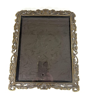 May include: A rectangular picture frame with an antique brass finish. The frame features an ornate border decorated with embossed floral motifs, leaves, and scrolling patterns. It contains a dark, reflective glass pane with a small circular hole.