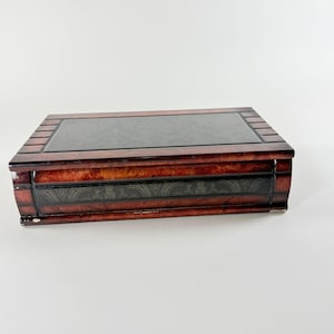 May include: A rectangular wooden box with a polished reddish-brown burl wood finish. The lid features a large, smooth, dark charcoal-colored inlay, while the front panel displays a horizontal band with a subtle, repeating gold-toned pattern.