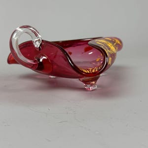 May include: A small, ruby red glass dish with a clear glass handle and three feet. The dish has a gold design on the side.