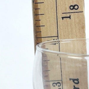 May include: A clear glass with a wooden ruler measuring 13 cm. The ruler is made of light brown wood with black markings. The glass is partially visible, with the text "Made in China" and "One Yard" printed on it.