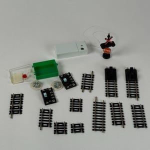 May include: A collection of miniature model train accessories, including track pieces, a crossing signal with a "STOP" sign, a white control box, and small green and clear plastic containers. The tracks are black with silver rails.
