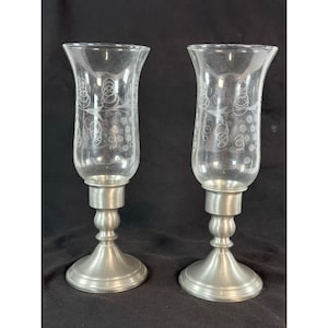 May include: Two clear glass candle holders with etched swirl designs. The glass tops sit on pewter-colored metal bases. The bases have a round foot, a central column, and a small platform to hold the glass. The candle holders are displayed against a black background.