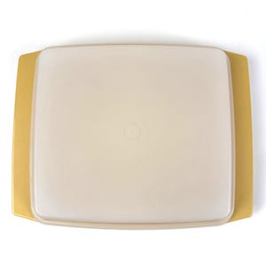 May include: A square, off-white plastic container with a matching lid. The lid has a slightly rounded shape and a small, circular indentation in the center. The container has a gold-colored base with extended edges.