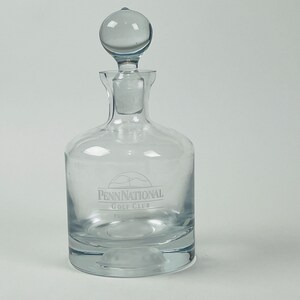 May include: A clear glass decanter with a round stopper. The decanter has a wide base and a narrow neck. The text "Penn National Golf Club" is etched on the front. The stopper is a clear, round ball.