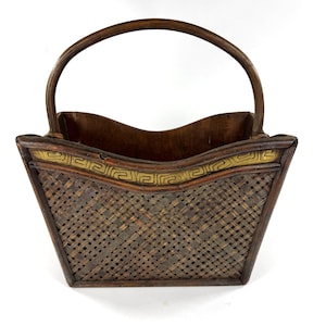 Peut inclure: Un panier tressé brun foncé avec une anse arquée unique. Le panier présente une ouverture large et évasée avec un bord supérieur ondulé. Une bande horizontale avec un motif grec doré est intégrée dans la partie supérieure du panier.