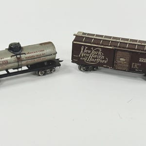 May include: Two vintage model train cars. One is a silver tank car with black wheels and the text "Niagara Chemicals Corp". The other is a brown boxcar with "New York, New Haven and Hartford" printed on the side.