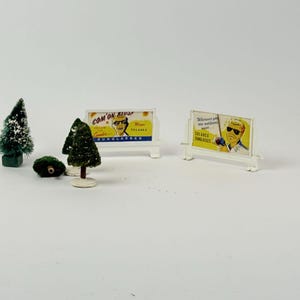 May include: Miniature vintage advertising billboards for Solarex sunglasses, featuring illustrations of a man wearing sunglasses. The billboards are white with yellow and blue text. Also, miniature green Christmas trees and a green bush are in the image.