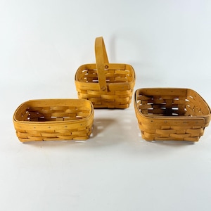 May include: Three light brown woven wooden baskets with a natural finish. One basket features a tall, upright handle, while the other two are open, rectangular containers. Each basket is constructed with horizontal and vertical wood strips.