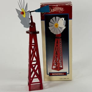 May include: A red metal windmill model with a gray fan and a blue tail vane, standing next to its original cardboard packaging box labeled Lemax Dickensvale Collectibles Windmill, featuring a graphic of the windmill on the front.