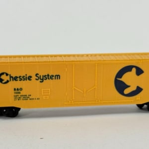 May include: A yellow model train boxcar with black wheels and accents. The words "Chessie System" are printed in black, along with a logo and other text. The boxcar is a miniature replica of a freight car.