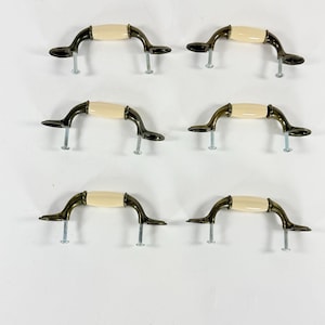 May include: Six antique-style cabinet handles. Each handle has a cream-colored center and dark bronze-colored curved arms. The handles are shown with the screws for installation. A classic hardware design.