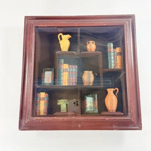 May include: A square, dark brown wooden shadow box display case with a glass front, containing miniature decorative items including small ceramic pitchers, stacks of books, ornate picture frames, and a classical column pedestal on internal shelves.