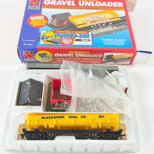 May include: A model train set featuring a yellow and black McKeepsport Coal Co. gondola car, a red side-dump car, and a black building. The box is labelled "Operating Gravel Unloader" and "HO Scale Scenery".