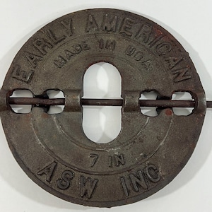 May include: A circular, cast iron object with the words "EARLY AMERICAN" and "MADE IN USA" embossed on the surface. The center has a large, elongated opening. The text "7 IN" is also visible. The object is from "ASW INC".