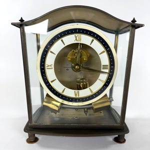 May include: An antique Junghans ATO clock with a white face, black and gold accents, and Roman numerals. The clock is encased in glass and has a brass pendulum and base. The clock is approximately 8 inches tall.