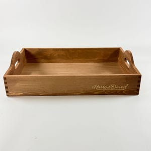 May include: A rectangular wooden tray with a natural finish. The tray has cut-out handles on each end and the words "Harry & David" are engraved on the front. The tray is made of wood and has a warm, brown color.