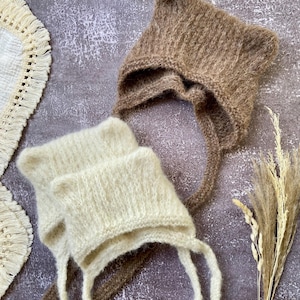 May include: Two knitted baby bonnets in the image. One is a light beige color, and the other is a medium brown. Both bonnets have ear-like points at the top and long ties. The bonnets are made of a soft, textured yarn.