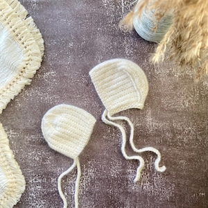 Peut inclure: Deux bonnets de bébé en crochet blanc avec des liens. Les bonnets sont sur une surface texturée brune.