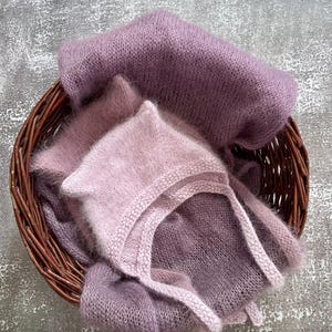 Puede incluir: Un conjunto de artículos de bebé tejidos y suaves en una cesta de mimbre. El conjunto incluye un gorro y una envoltura en tonos lavanda y malva. Los artículos están hechos de un material delicado y difuso, ideal para fotografía de recién nacidos.