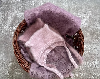 Puede incluir: Un conjunto de artículos de bebé tejidos y suaves en una cesta de mimbre. El conjunto incluye un gorro y una envoltura en tonos lavanda y malva. Los artículos están hechos de un material delicado y difuso, ideal para fotografía de recién nacidos.