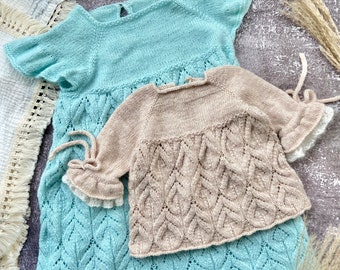 May include: Two knitted baby dresses, one in light blue and one in beige, with a leaf pattern. The dresses have ruffled sleeves and are laid out on a textured surface.