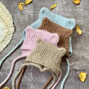 May include: Four knitted baby bonnets in pastel colors: light blue, brown, pink, and beige. Each bonnet has ear-like points and long ties. The bonnets are arranged on a textured gray surface with scattered yellow petals.