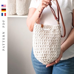 Może przedstawiać: Kremowa, szydełkowa torba na ramię z brązowym skórzanym paskiem i akcentami. Torba jest trzymana przez osobę ubraną w białą koszulę i niebieskie dżinsy. Dwie podobne torby znajdują się na półce w tle. Obraz zawiera tekst "PATTERN PDF".