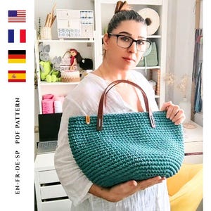 May include: Teal crochet tote bag with brown leather handles. The bag is held by a person. The text "EN-FR-DE-SP PDF PATTERN" is visible on the left side of the image.