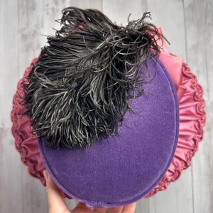 May include: A purple velvet hat with a black feather on top. The hat is trimmed with a pink satin ruffle.