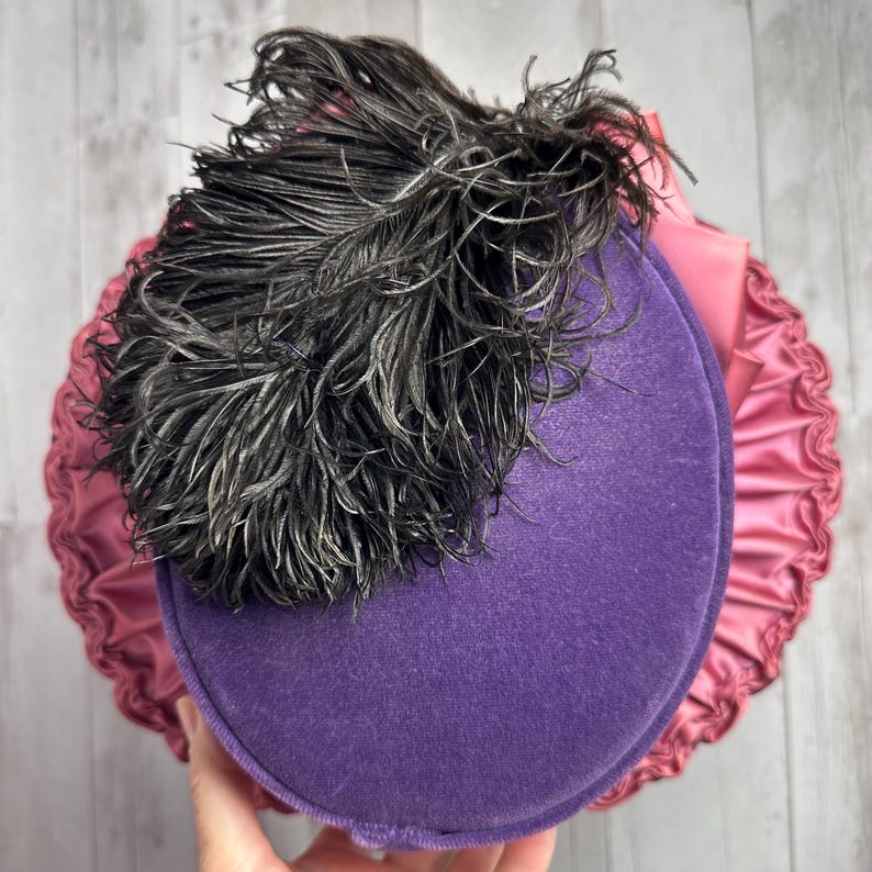 May include: A purple velvet hat with a black feather on top. The hat is trimmed with a pink satin ruffle.