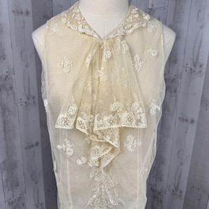 Puede incluir: Una blusa sin mangas de color crema con intrincados detalles de encaje. La blusa presenta un frente con volantes y un delicado cuello de encaje. El encaje está adornado con patrones florales, creando una estética vintage. La blusa se exhibe en un maniquí.