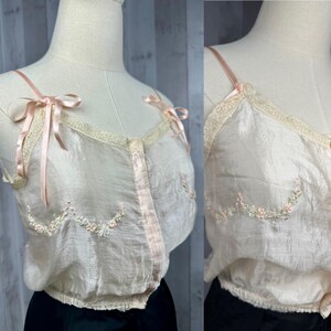 May include: A delicate, sheer, blush-colored camisole with delicate floral embroidery and satin ribbon straps. The camisole has a button-up front and a gathered hem.
