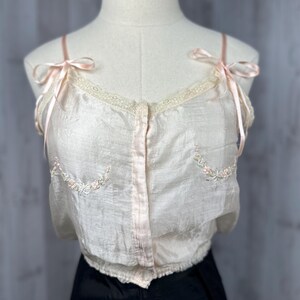 May include: A delicate, off-white silk camisole with pink satin straps and floral embroidery. The camisole has a button-up front and a lace trim around the neckline.