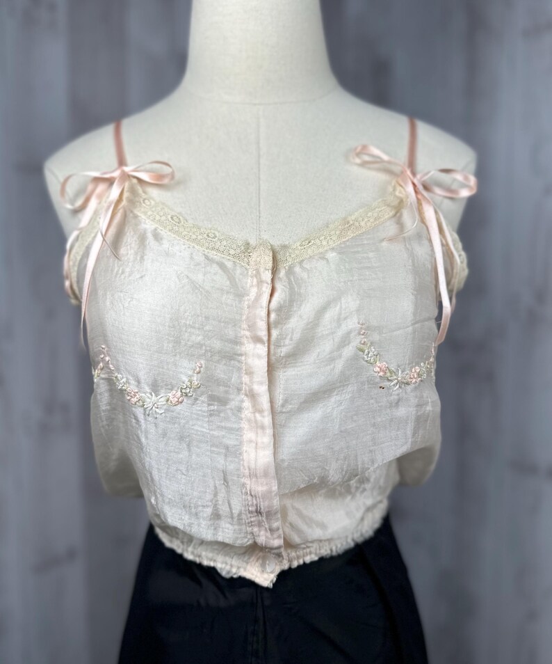 May include: A delicate, off-white silk camisole with pink satin straps and floral embroidery. The camisole has a button-up front and a lace trim around the neckline.