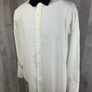Pu&ograve; includere: Una camicia bianca a maniche lunghe con un papillon nero. La camicia ha una chiusura con bottoni sul davanti e un colletto a punta.