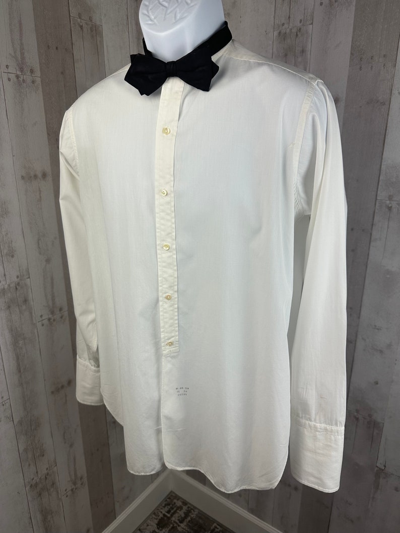 Pu&ograve; includere: Una camicia bianca a maniche lunghe con un papillon nero. La camicia ha una chiusura con bottoni sul davanti e un colletto a punta.