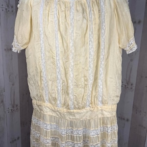 May include: A pale yellow blouse featuring short sleeves, a high neckline with white lace trim, and vertical white lace panels running down the front. The garment includes a gathered waistline and a lower section detailed with horizontal white lace.