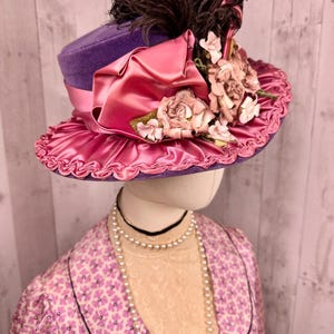 May include: A purple velvet hat with a pink satin ribbon and flower embellishments. The hat has a brown feather on top.