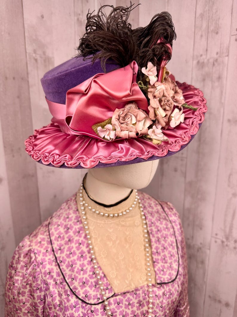 May include: A purple velvet hat with a pink satin ribbon and flower embellishments. The hat has a brown feather on top.