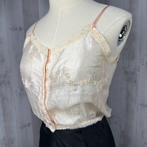 May include: A pale pink satin camisole with delicate floral embroidery and lace trim. The camisole has a button-up front and thin straps.