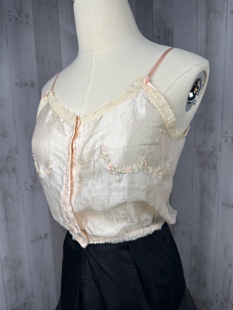May include: A pale pink satin camisole with delicate floral embroidery and lace trim. The camisole has a button-up front and thin straps.
