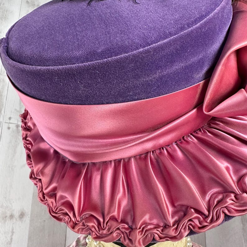 May include: A purple velvet hat with a pink satin ribbon and ruffle trim.