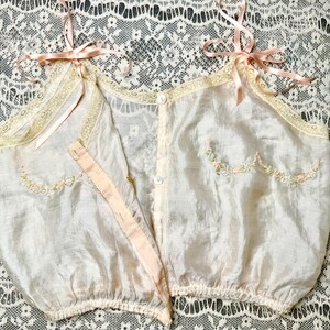 May include: A delicate, pale pink silk camisole with embroidered floral details and lace trim. The camisole has a button closure at the front and adjustable straps.
