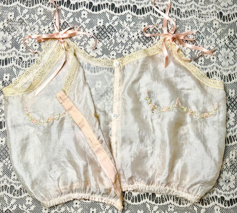 May include: A delicate, pale pink silk camisole with embroidered floral details and lace trim. The camisole has a button closure at the front and adjustable straps.