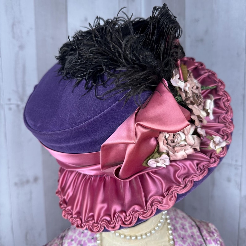 May include: A purple velvet hat with a pink satin ruffle and a black feather trim. The hat is decorated with pink fabric flowers.