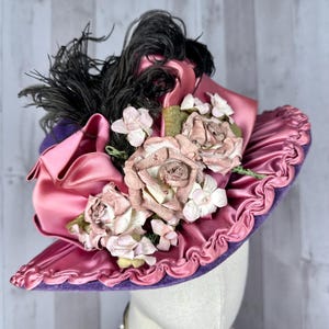 May include: A vintage hat with a wide brim, made of purple velvet and pink satin. The hat is decorated with pink fabric roses, white flowers, and black feathers.