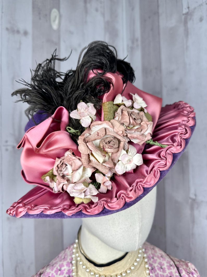 May include: A vintage hat with a wide brim, made of purple velvet and pink satin. The hat is decorated with pink fabric roses, white flowers, and black feathers.